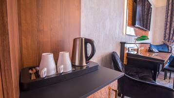 Una habitación de hotel con una pared de madera decorativa, una encimera negra con una tetera y dos tazas blancas, una silla y una mesa con una lámpara verde.