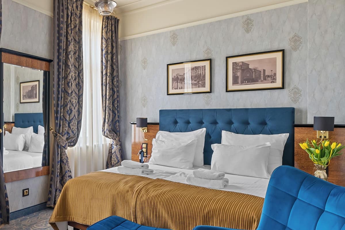 A hotel room with a large bed, a blue headboard, a mirror, two framed pictures on the wall, and a vase with flowers.