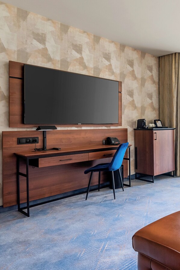 A hotel room with a large flat-screen TV mounted on the wall, a wooden desk with a chair, a small cabinet, and a leather ottoman.
