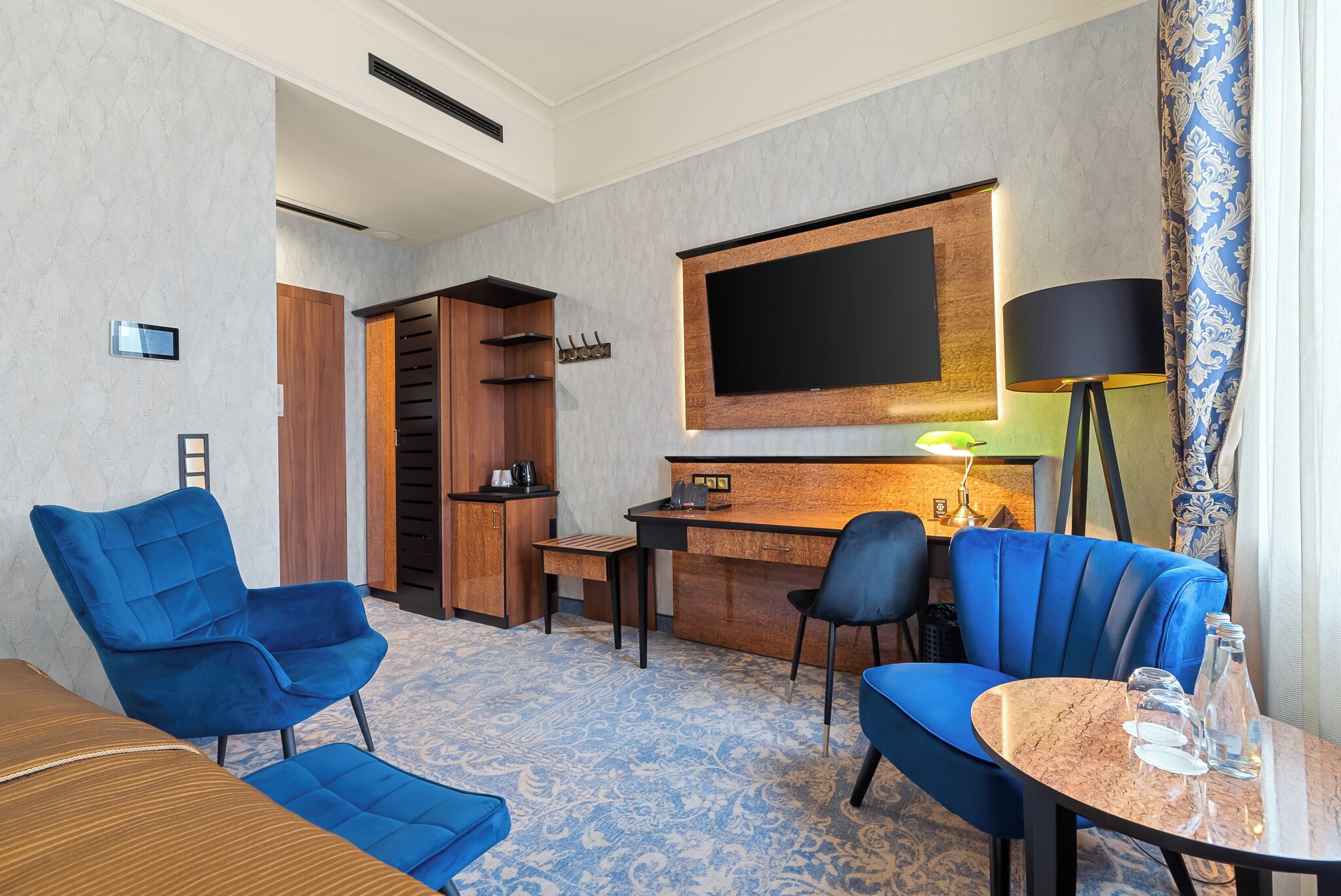Ein Hotelzimmer mit einem Bett, zwei blauen Sesseln, einem kleinen runden Tisch, einem Schreibtisch, einem Fernseher und einer Lampe.
