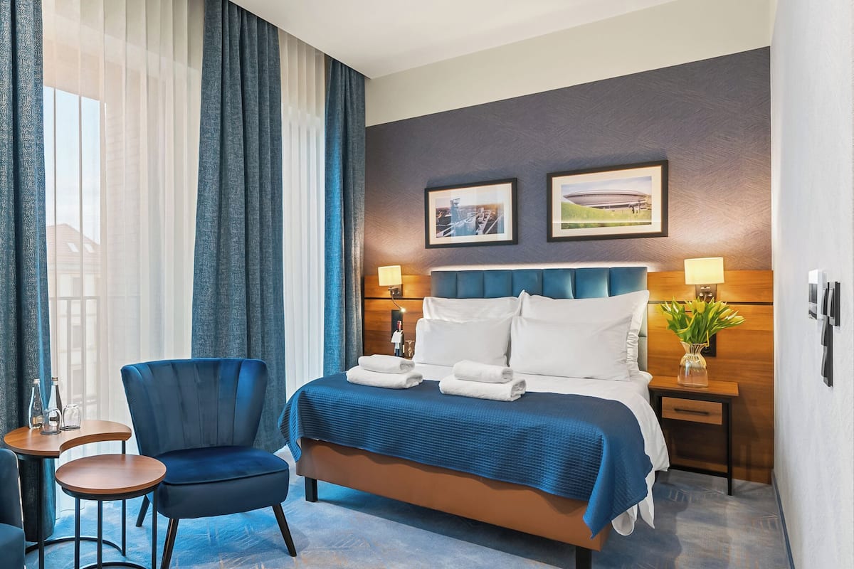 A hotel room with a bed, a chair, a small table, and a nightstand with a lamp.