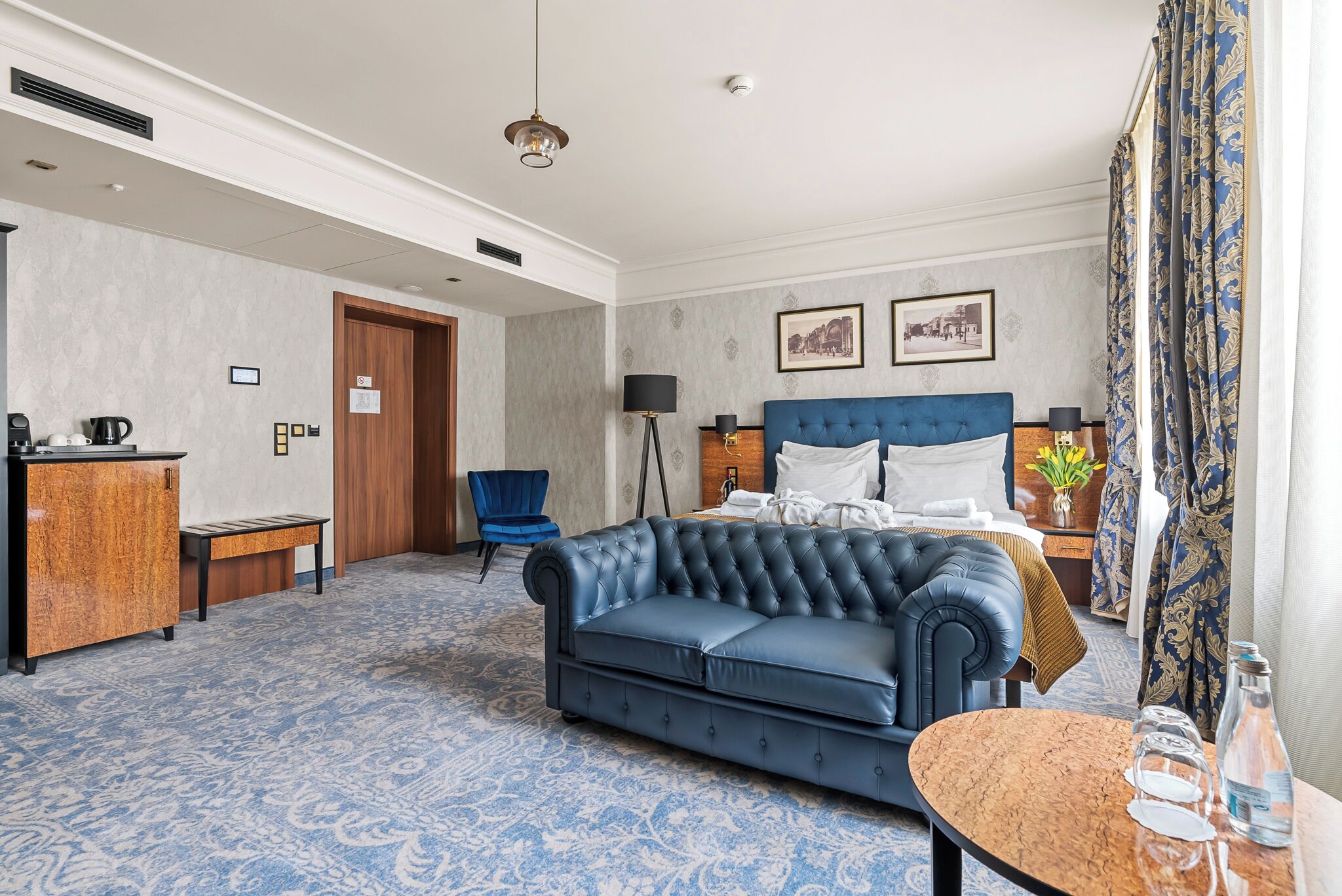 A hotel room with a large bed, a blue sofa, a small table, a desk, a chair, and a window with curtains.
