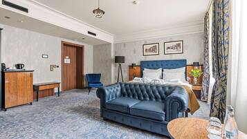 A hotel room with a large bed, a blue sofa, a small table, a desk, a chair, and a window with curtains.