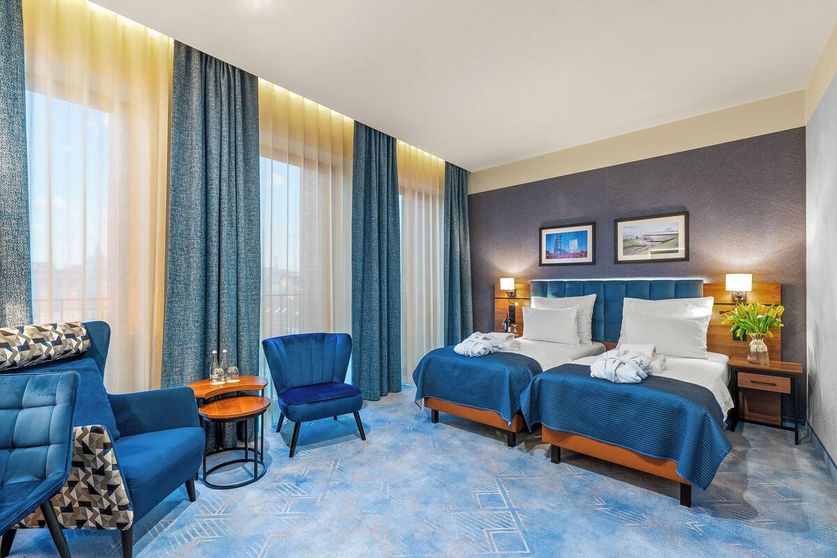 A hotel room with two beds, a sofa, a chair, a small table, and a nightstand with a lamp.