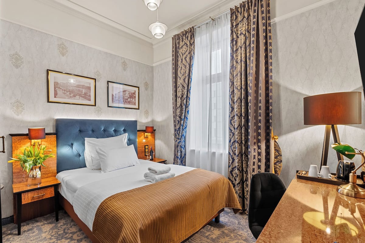 A hotel room with a large bed, a desk, a chair, and a nightstand with a lamp.