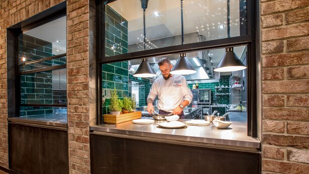 Un chef preparando comida en una cocina con paredes de ladrillo y ventanas amplias.