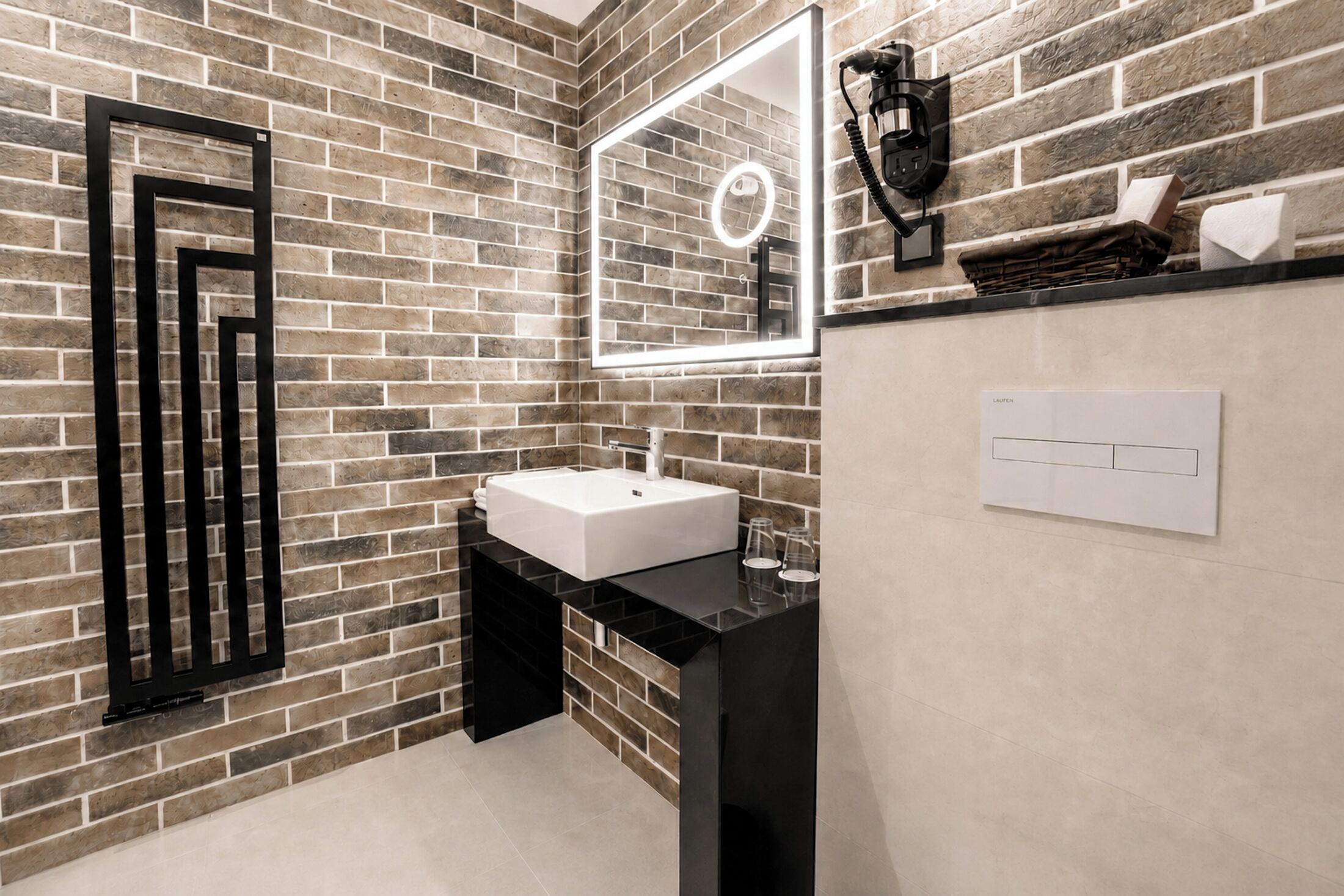 Un baño con pared de ladrillo, un lavabo blanco, un mueble de tocador negro, un espejo con luz y un perchero para toallas.