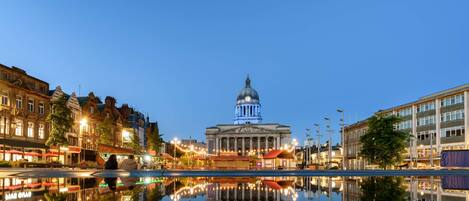 A historic building with a dome, reflecting in a calm water body, surrounded by urban buildings and street lights.