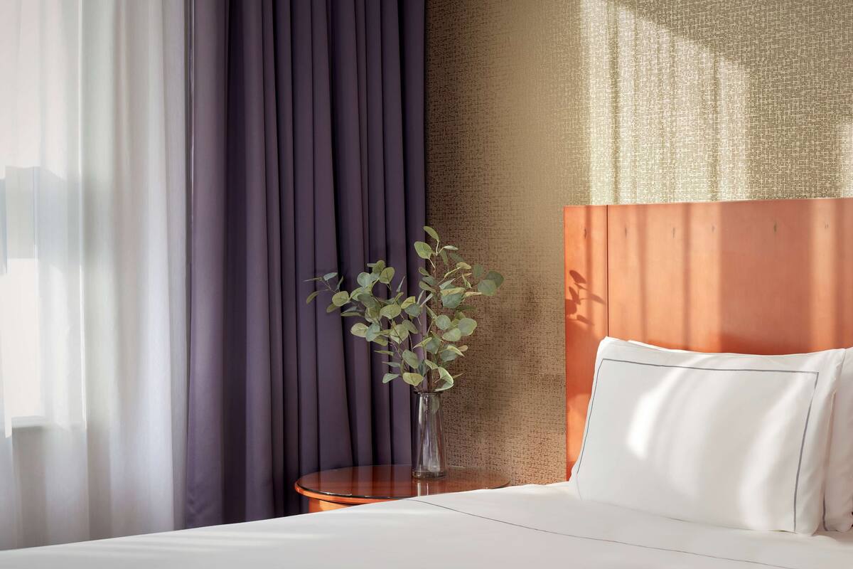 Ein Hotelzimmer mit einem Bett, einem kleinen runden Tisch, einer Vase mit Pflanze und einer strukturierten Wand.
