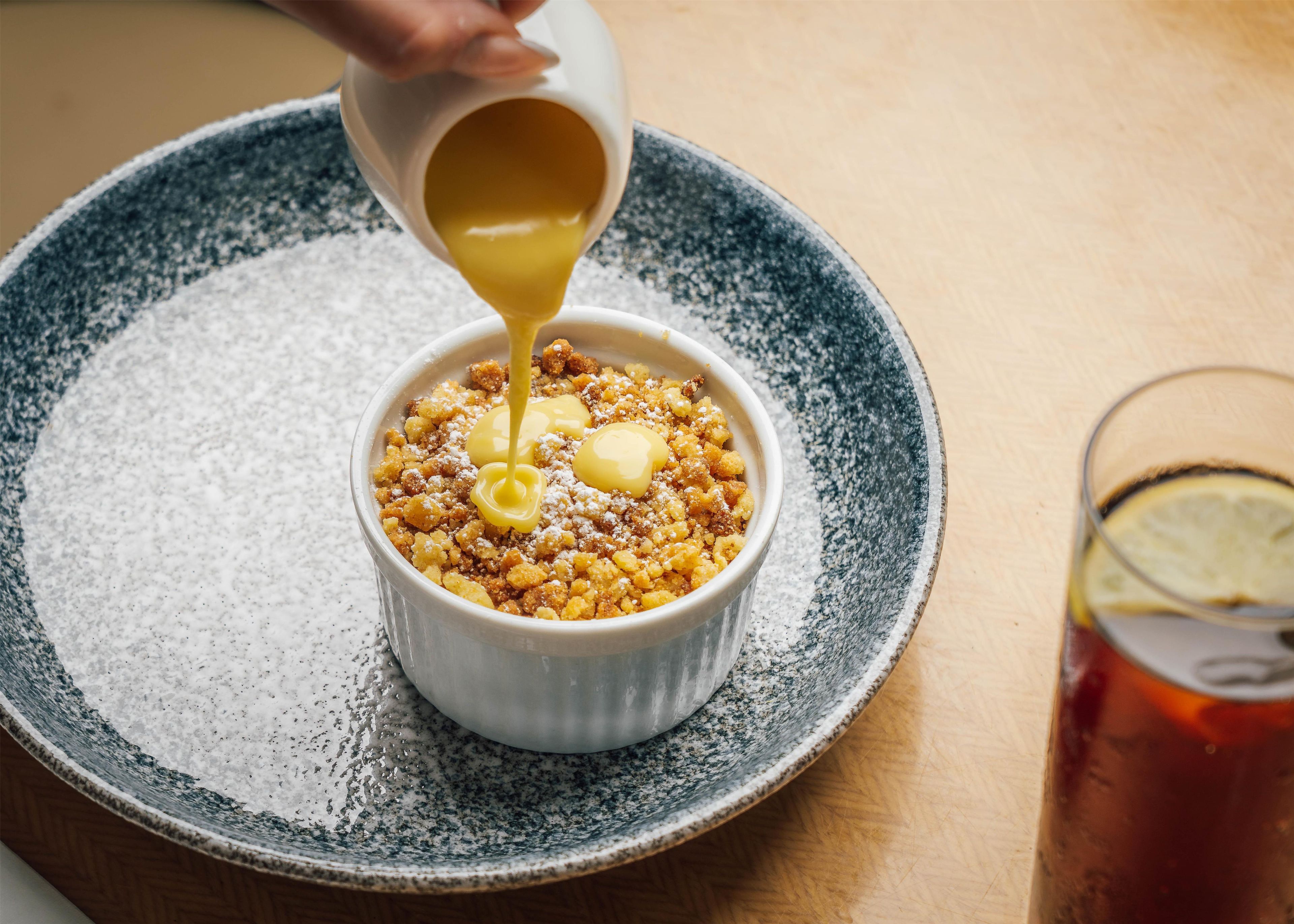 A dessert in a ceramic dish on a speckled plate, with a hand pouring a yellow liquid over it.