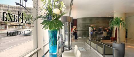 A modern hotel lobby with a reception desk, seating area, and a large window with a view of the street.
