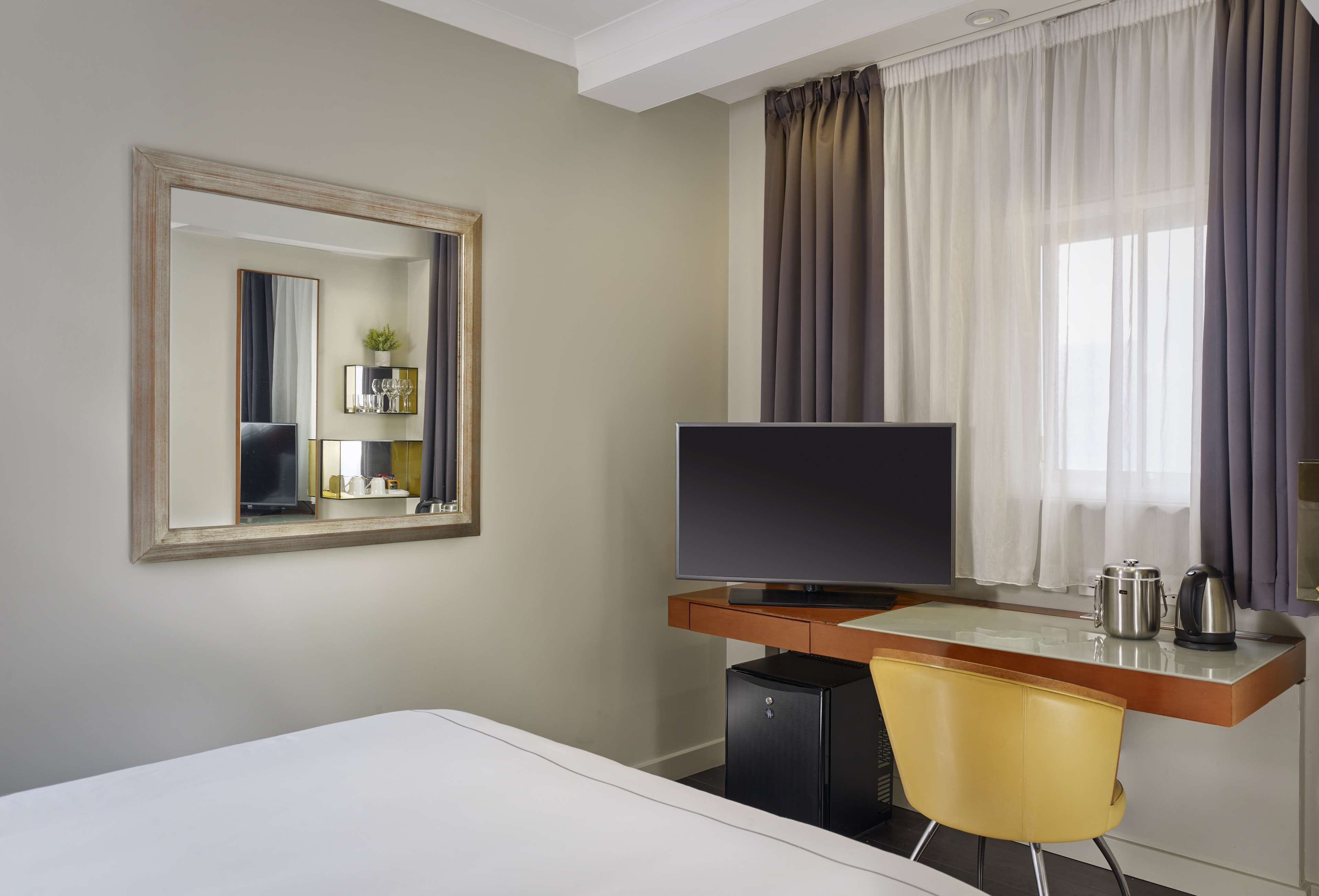 Quarto de hotel com uma cama, escrivaninha com computador, televisão, espelho e janela com cortinas.