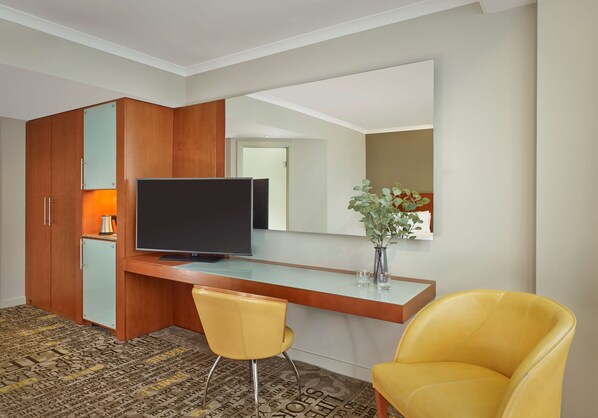 A hotel room with a wooden wardrobe, a flat-screen TV, a small fridge, a yellow armchair, and a patterned carpet.