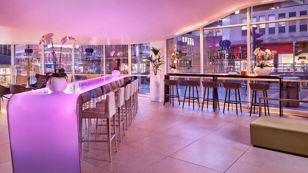 Un área de bar moderna con una barra larga, varios taburetes y grandes ventanales con vistas a la ciudad.