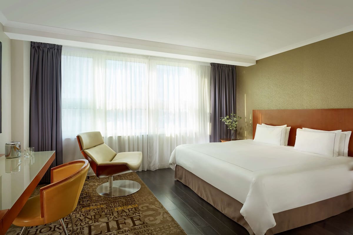 Ein Hotelzimmer mit einem großen Bett, einem Schreibtisch mit Stuhl, einem kleinen Tisch und einem großen Fenster mit Vorhängen.
