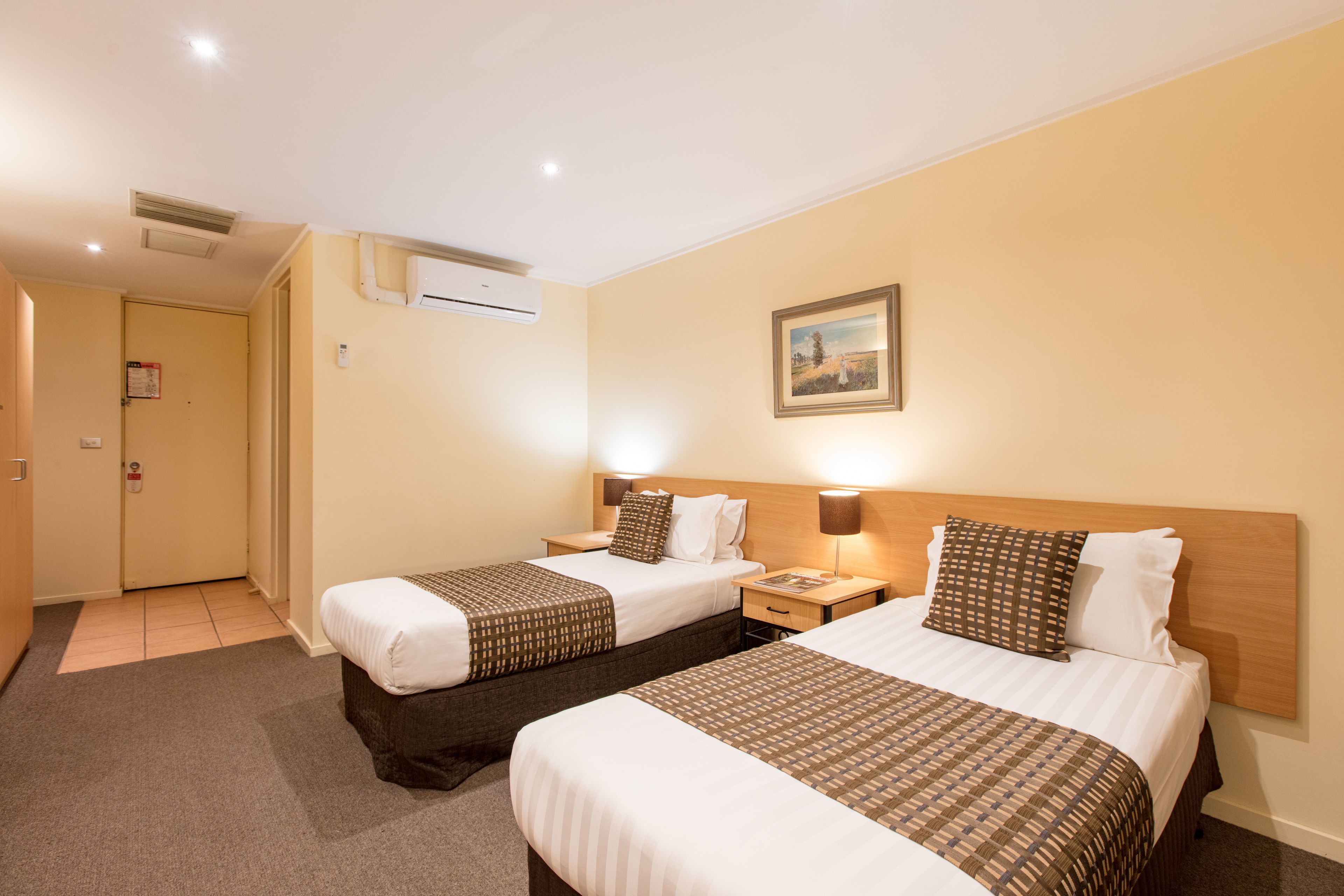 Une chambre d’hôtel avec deux lits, une table de chevet, un climatiseur fixé au mur et un tableau encadré accroché au mur.