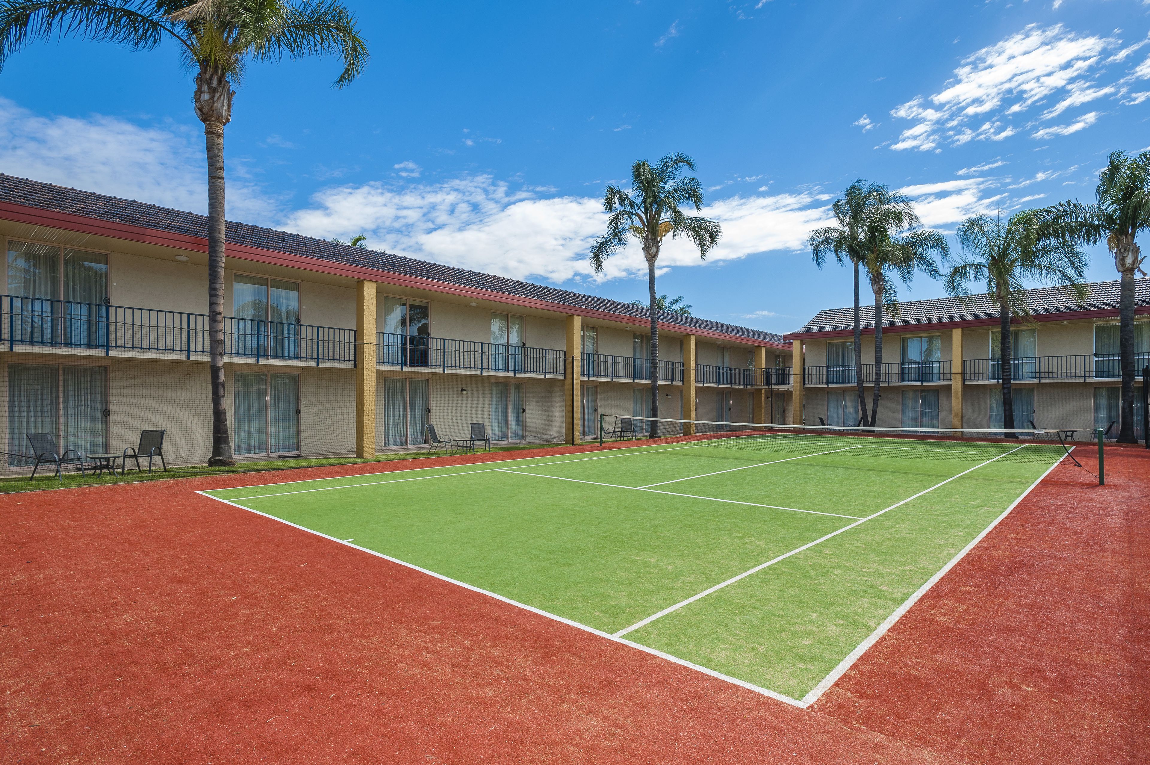 A tennis court with a net, surrounded by a building with balconies and palm trees.