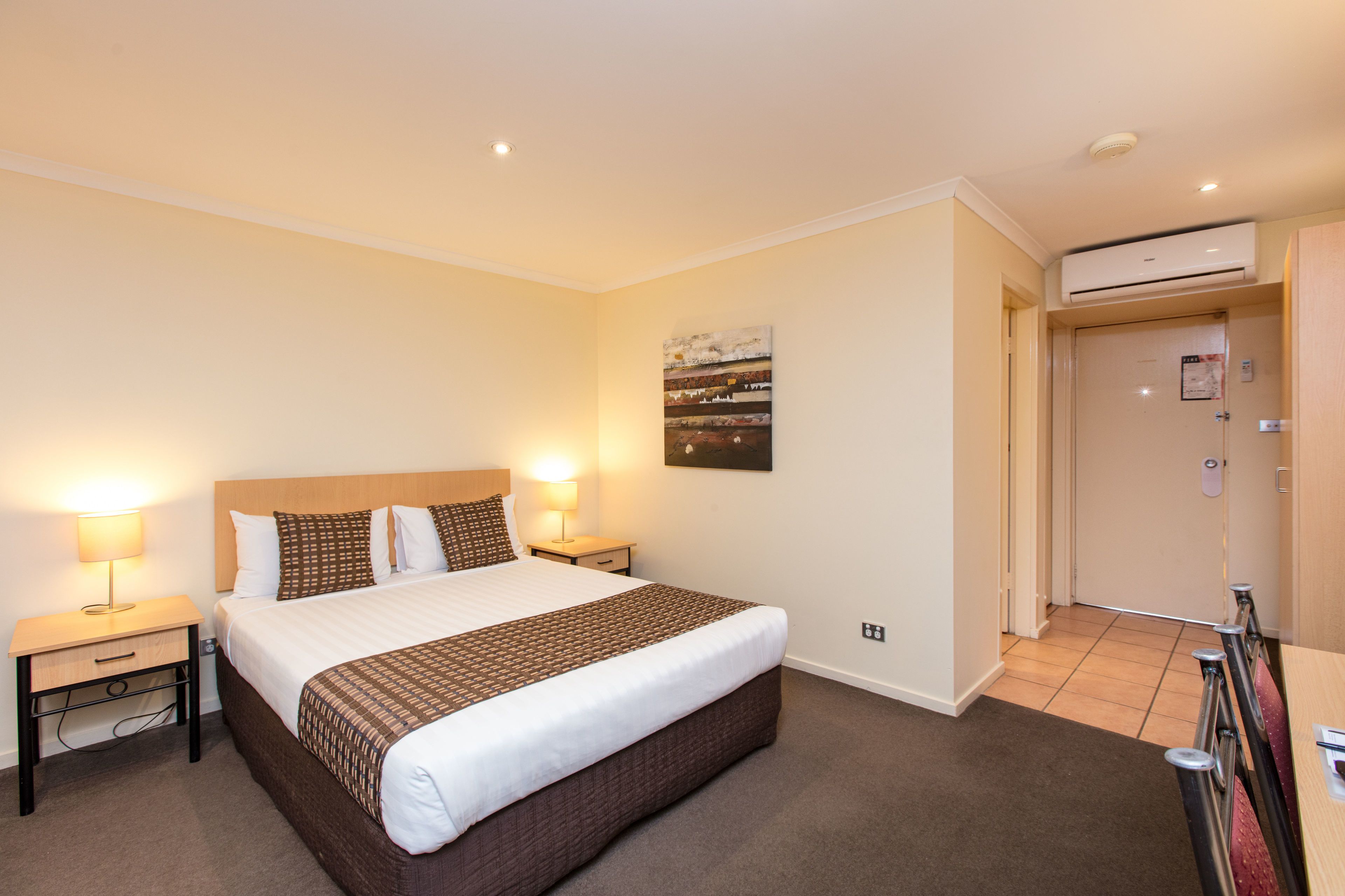 A hotel room with a large bed, bedside tables with lamps, a wall-mounted air conditioner, and a door leading to another room.