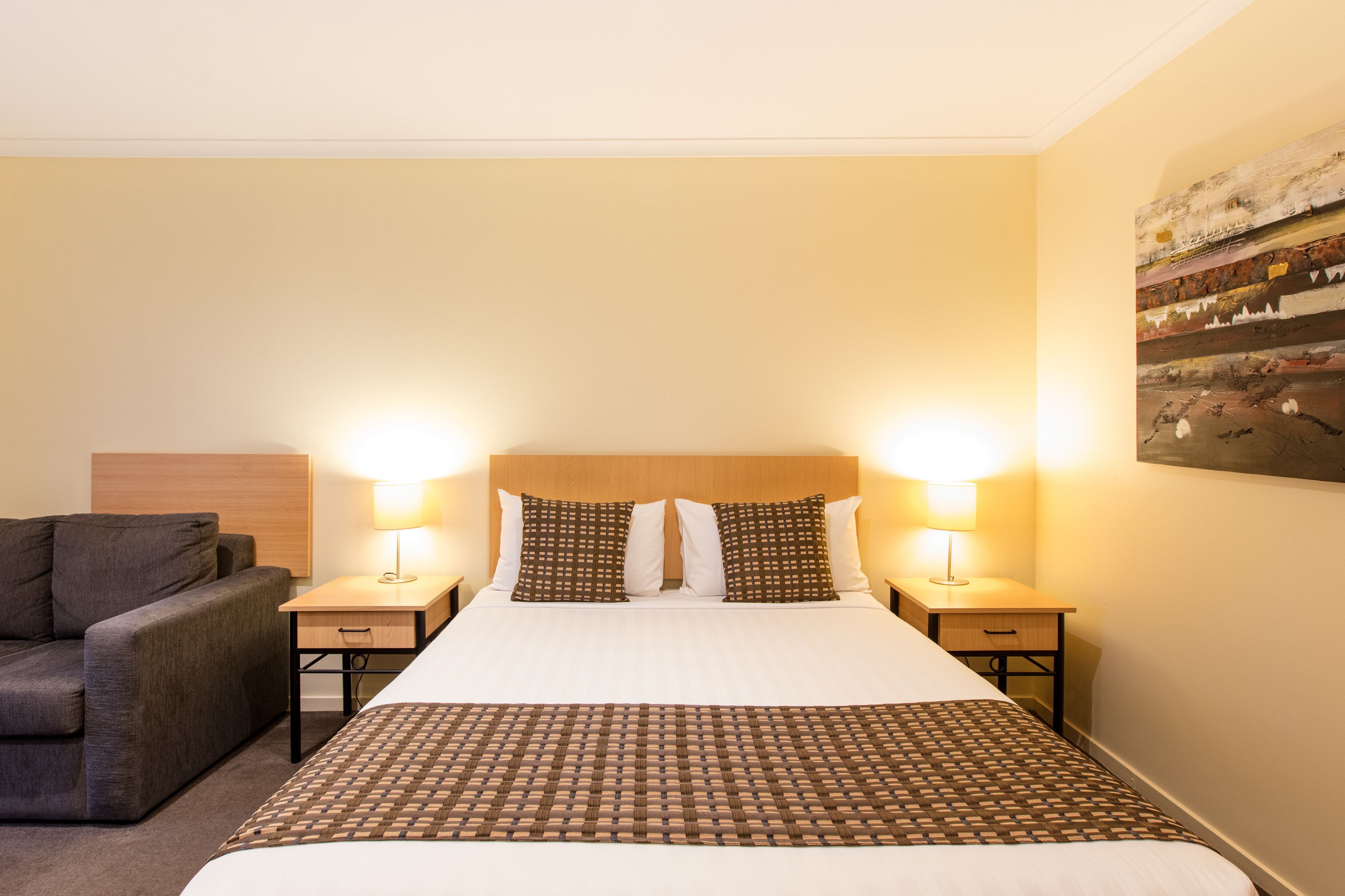 A hotel room with a large bed, two bedside lamps, a grey sofa, and a wall-mounted artwork.