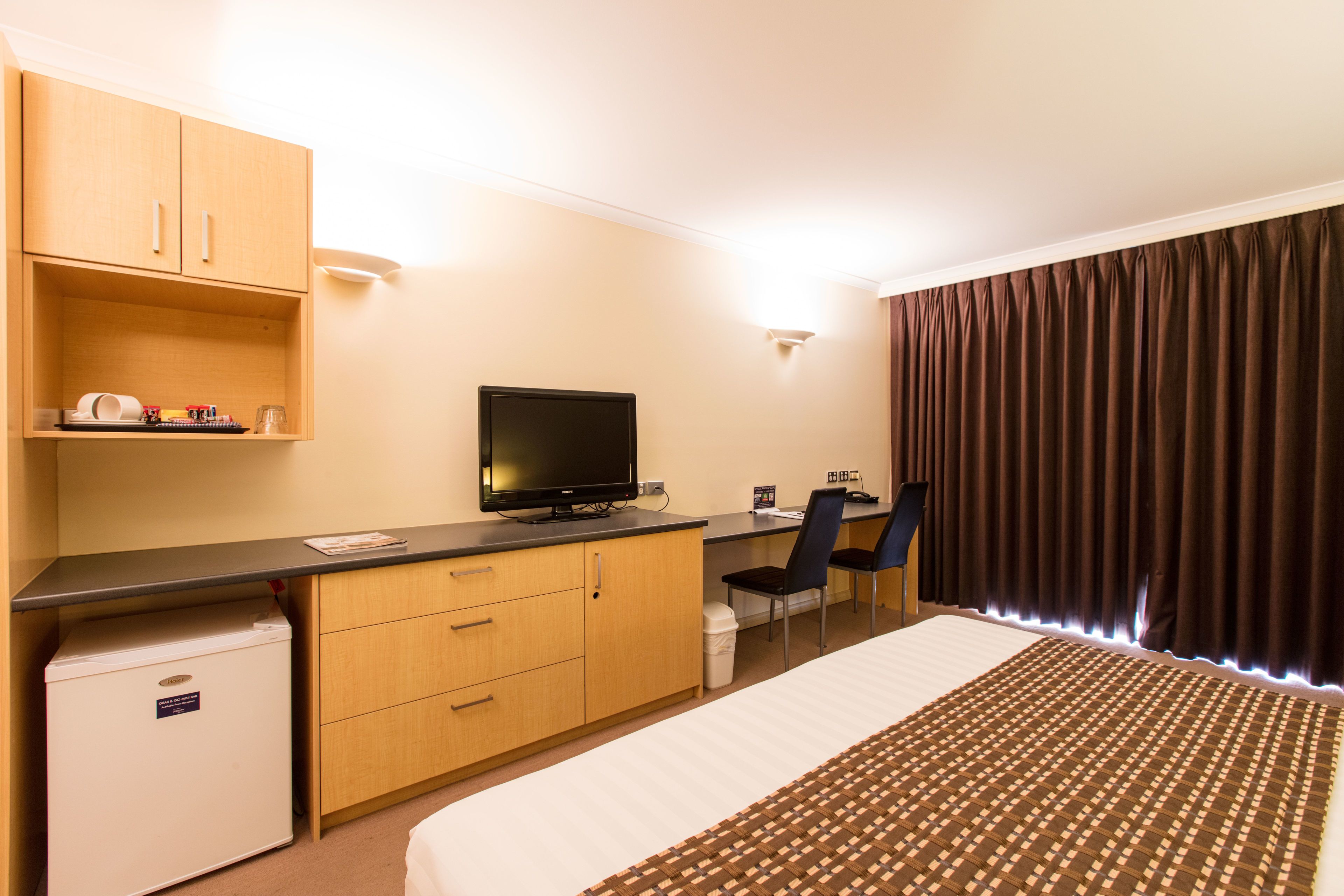 A hotel room with a bed, a desk with a computer, a television, a chair, and a small refrigerator.