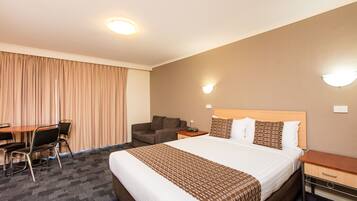 Quarto de hotel com uma cama grande, uma mesa com cadeiras, um sofá e duas luminárias fixadas na parede.