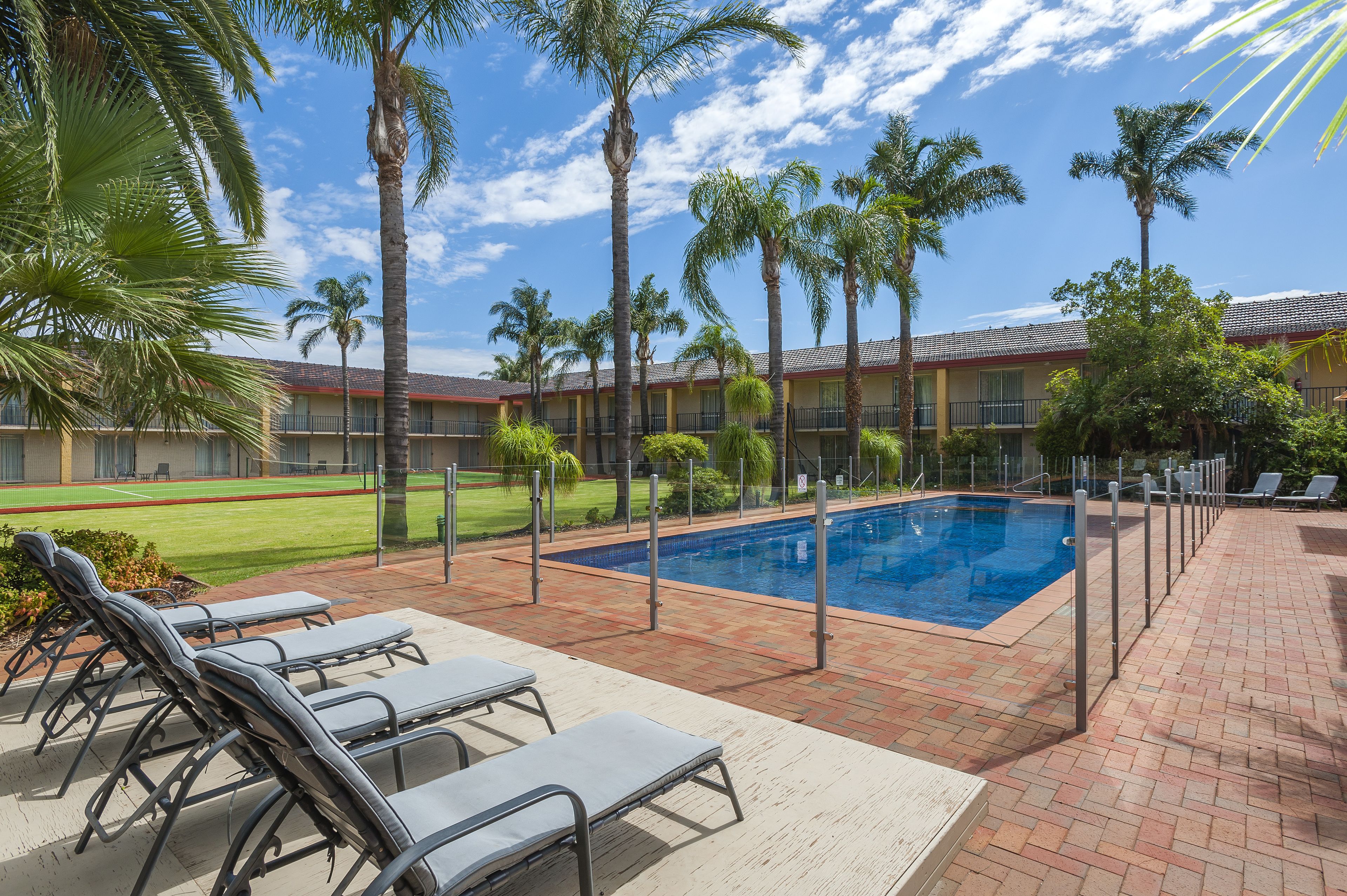 A poolside area with lounge chairs, a swimming pool, palm trees, and a building with a red roof in the background.