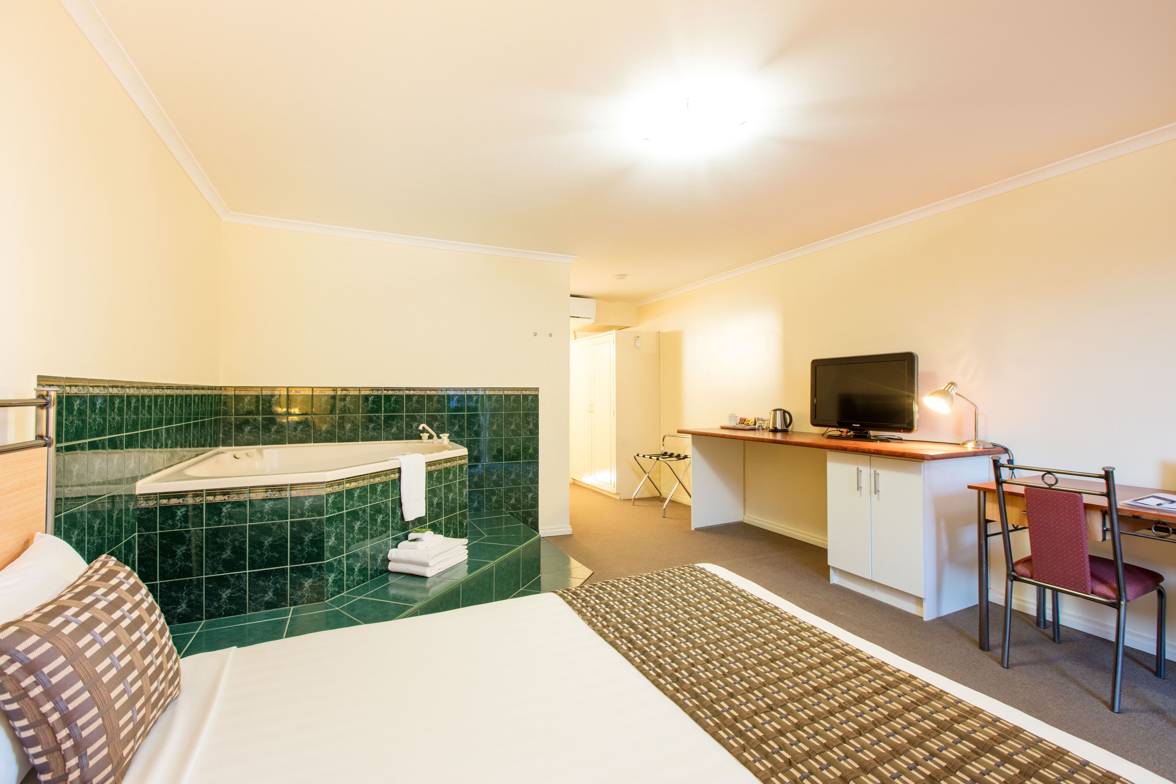 Una habitación de hotel con una cama, un escritorio con una silla, un televisor y un baño con bañera y azulejos verdes.