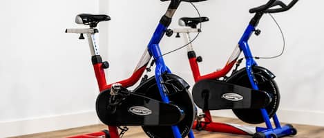 To stillestående cykler med røde og blå stel, sorte sadler og et sort svinghjul, placeret på et trægulv foran en hvid væg.