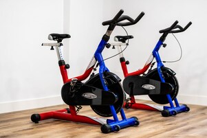 Två motionscyklar med röda och blå ramar, svarta sadlar och ett svart svänghjul, placerade på ett trägolv framför en vit vägg.