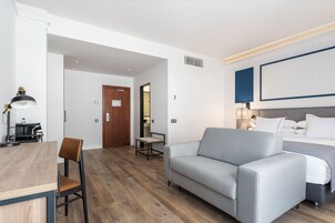 A modern hotel room with a grey sofa, a wooden dining table with two chairs, a small desk with a lamp, and a bed with white bedding.