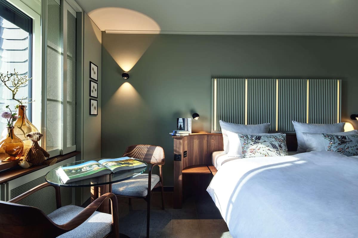 Ein Hotelzimmer mit einem großen Bett, einem Nachttisch mit Lampe, einem Sessel, einem kleinen Tisch mit einem Buch und einem Fenster mit Jalousien.