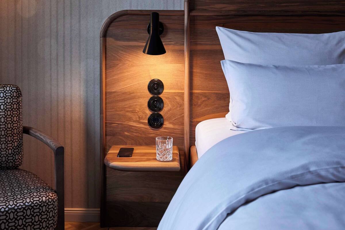 Ein ordentlich eingerichtetes Hotelzimmer mit einem hölzernen Kopfteil, einem Nachttisch mit Lampe, einem gemusterten Sessel und einem Bett mit weißen Leinen.