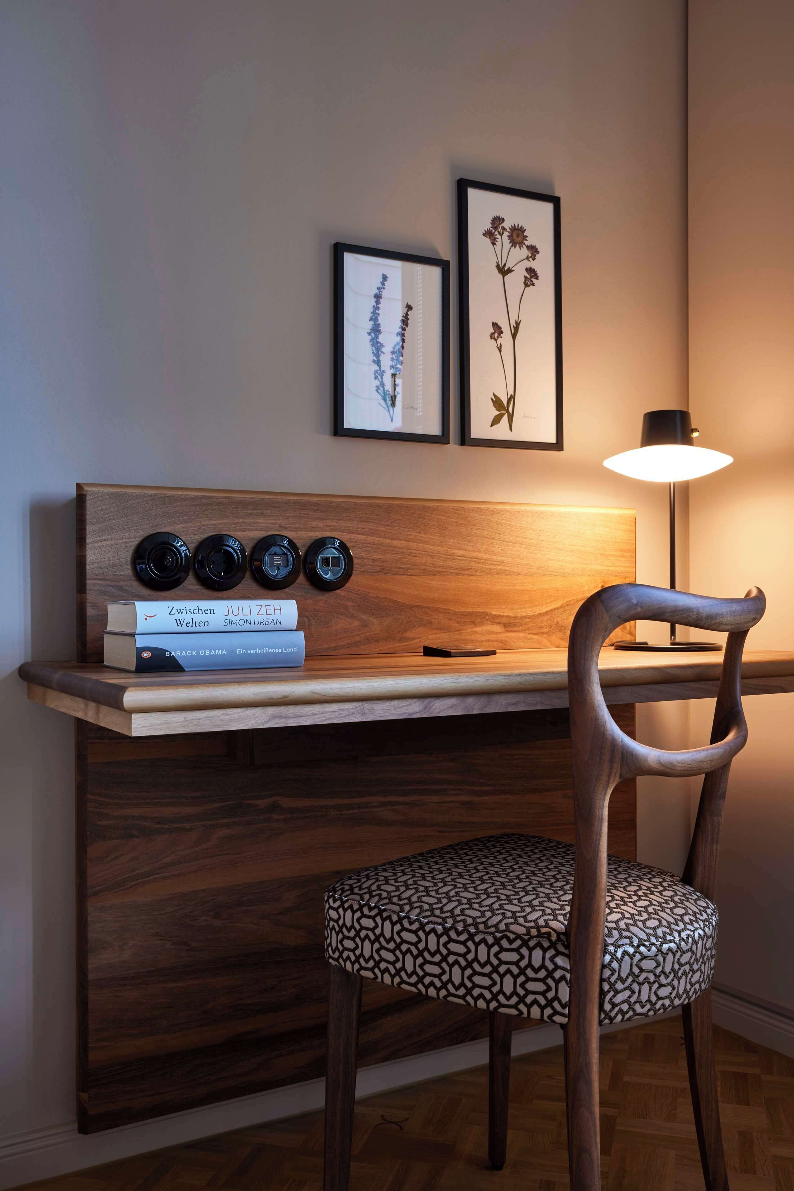 A modern desk with a wooden finish, a chair with a patterned cushion, and two framed botanical prints on the wall.