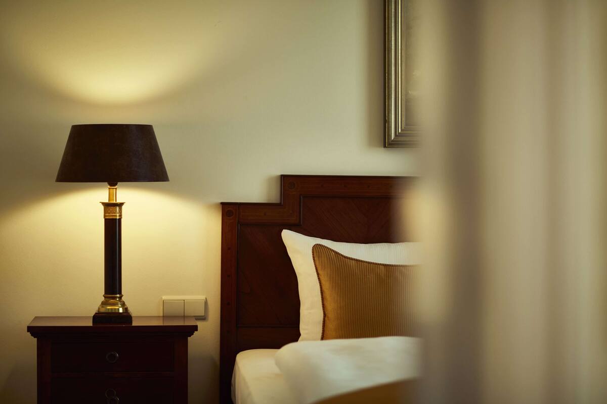 Ein Hotelzimmer mit Nachttisch aus Holz, einer Lampe, einem Bett mit weißer und goldener Bettwäsche und einem gerahmten Bild an der Wand.