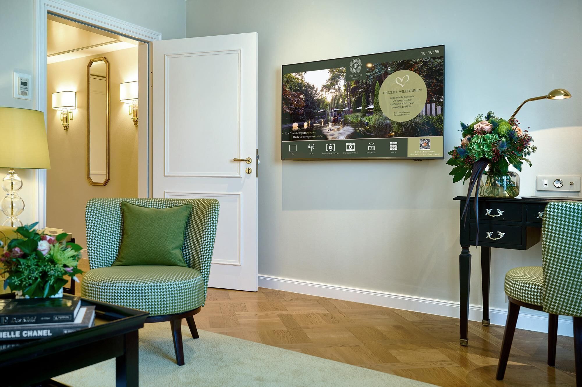Un salón bien iluminado con un sillón verde estampado, una mesa de centro negra con libros, y un televisor de pared que muestra un mensaje.