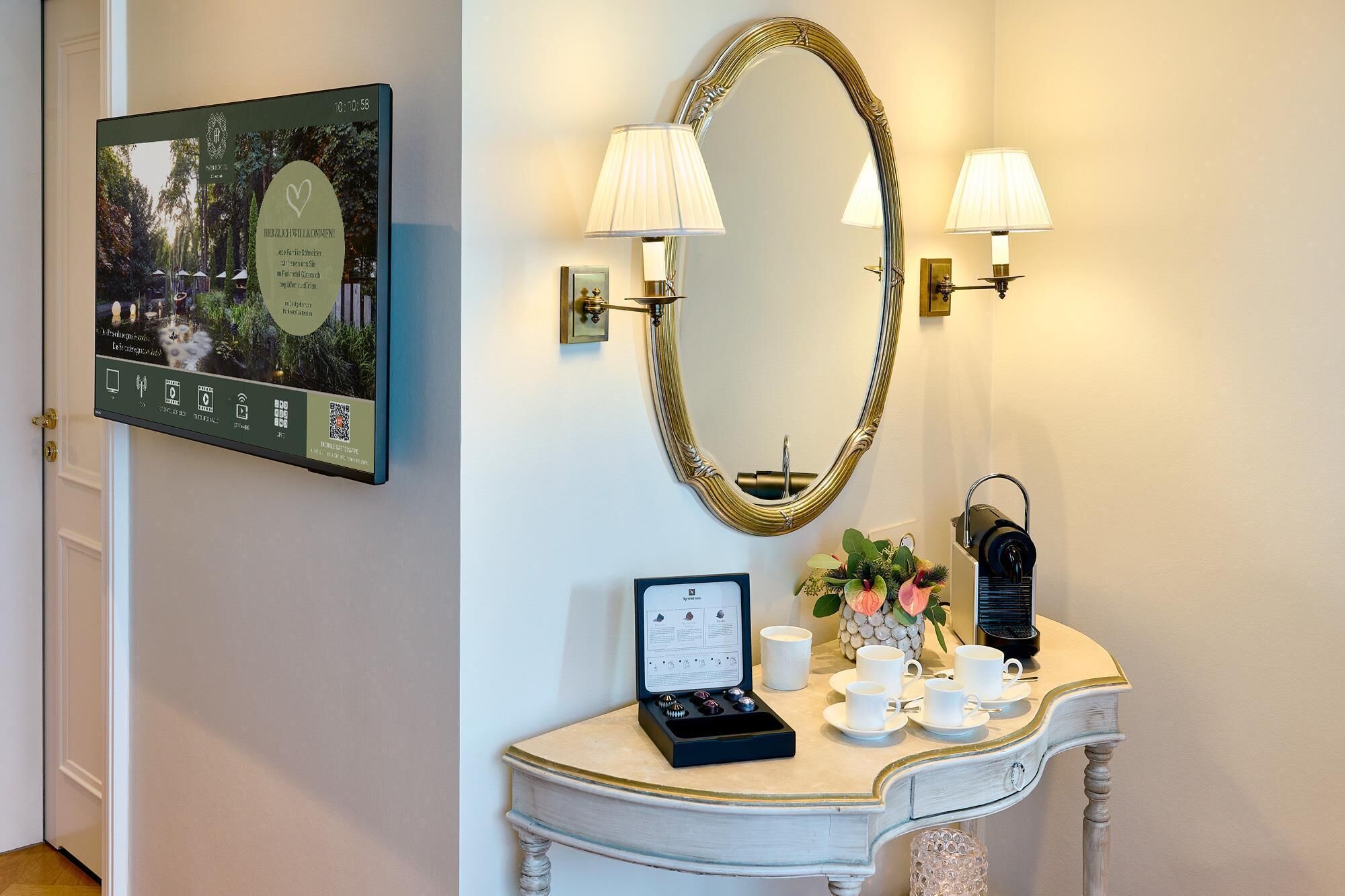 A room with a framed picture of a garden, a gold-framed mirror, two wall-mounted lamps, a small table with a tea set, and a decorative vase.