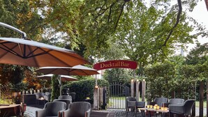 An outdoor seating area with wicker chairs and tables, a red sign for Duck Rail Bar, and a cobblestone floor. - Parkhotel Gütersloh (Guetersloh)