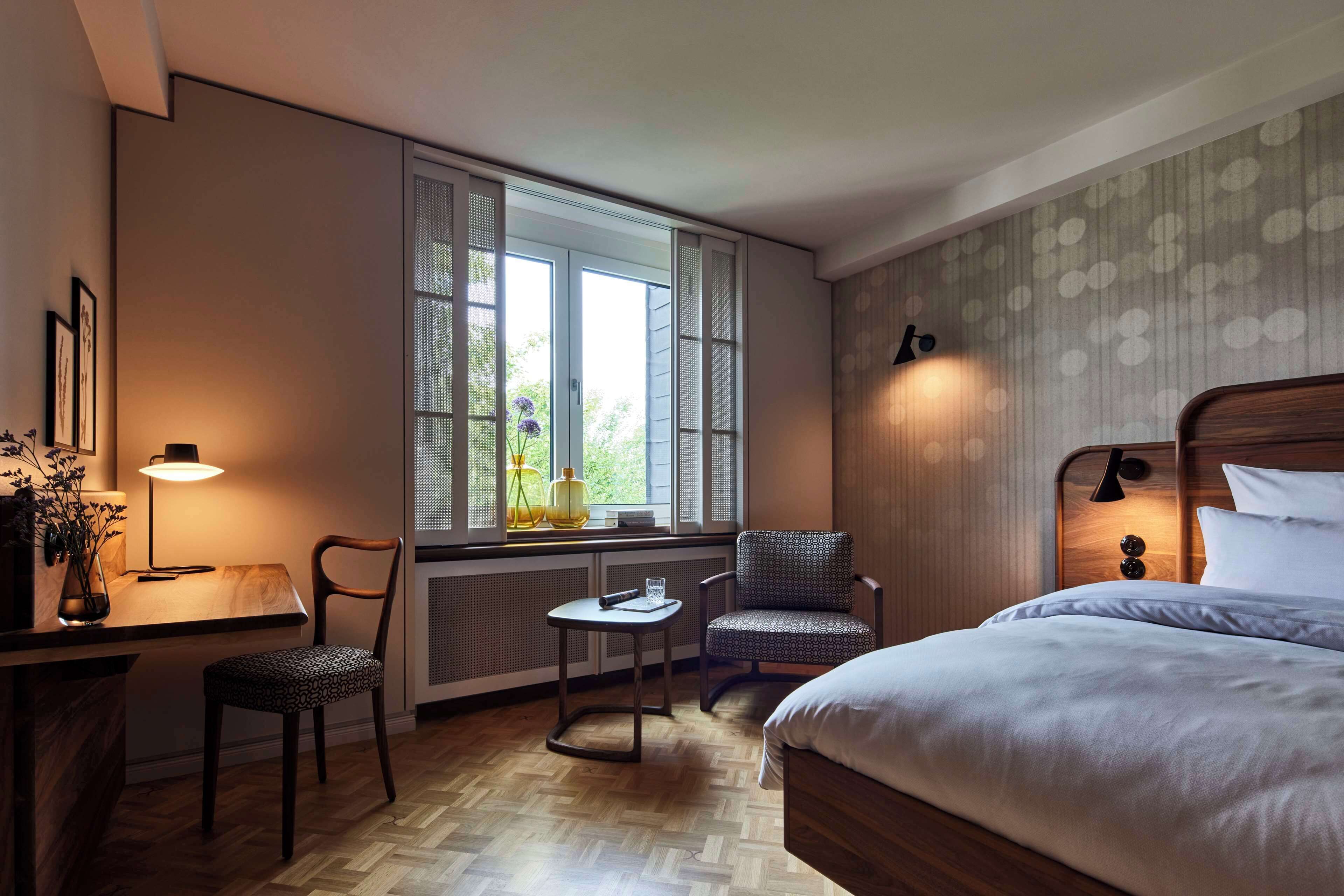 A hotel room with a bed, a desk with a lamp, a chair, a small table, and a window with shutters.