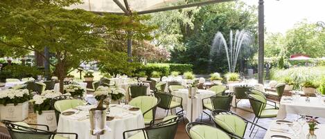 An outdoor dining area with round tables, green cushioned chairs, and white tablecloths.