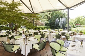 An outdoor dining area with round tables, green cushioned chairs, and white tablecloths.