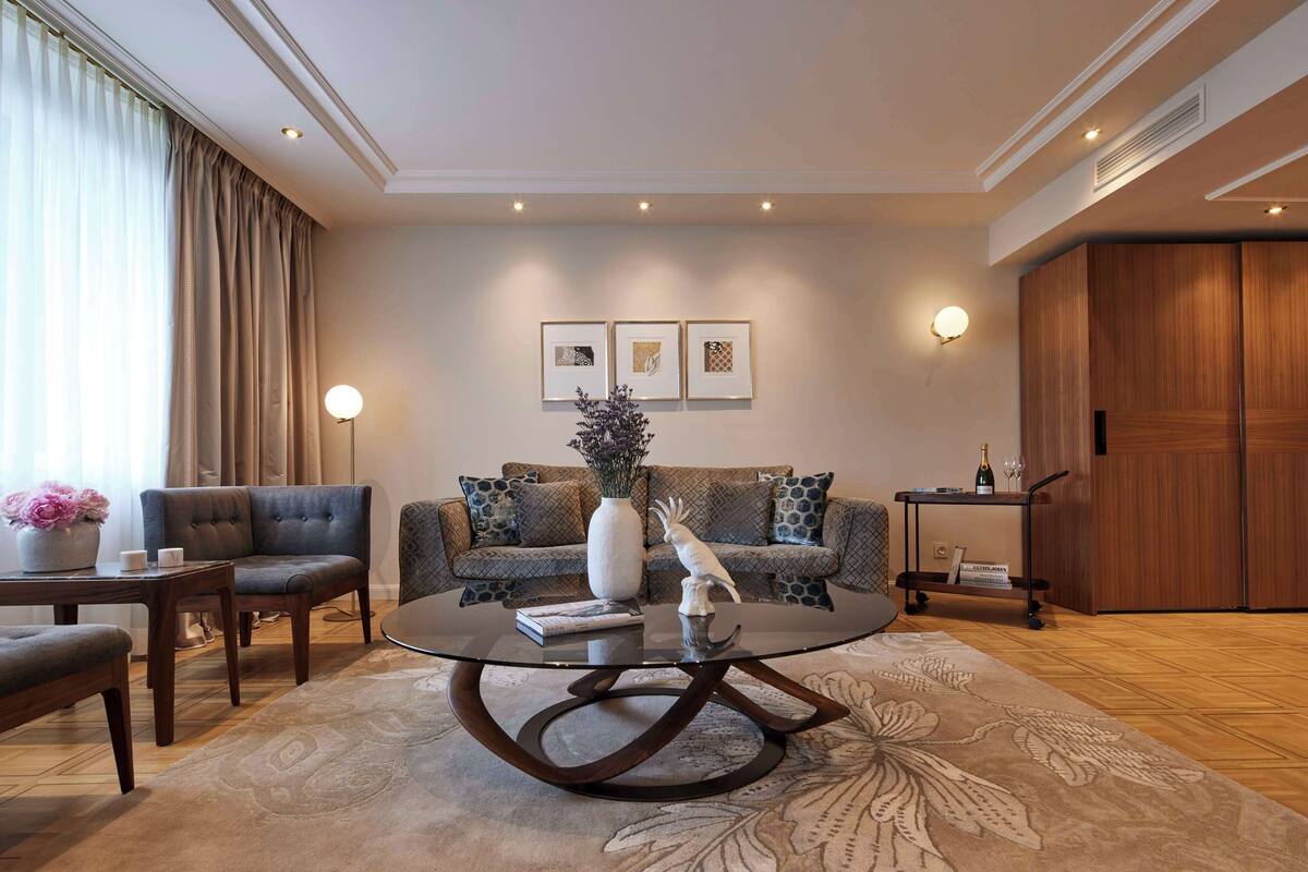 Ein modernes Wohnzimmer mit einem Glastisch, einer Couch, einem Sessel, einem Beistelltisch, einer Vase mit Blumen und einem gerahmten Bild an der Wand.