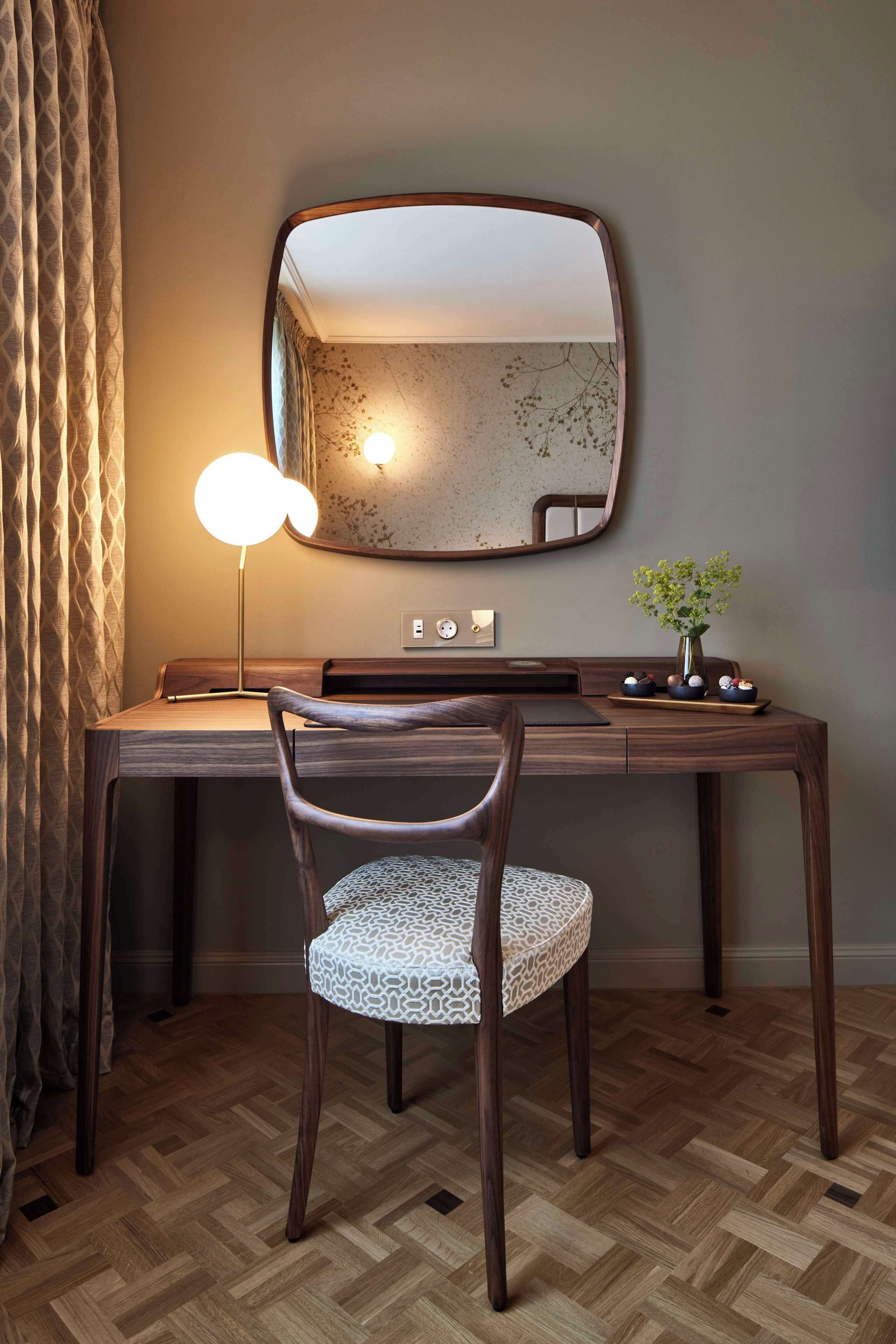 A wooden desk with a mirror, a desk lamp, and a vase with flowers.