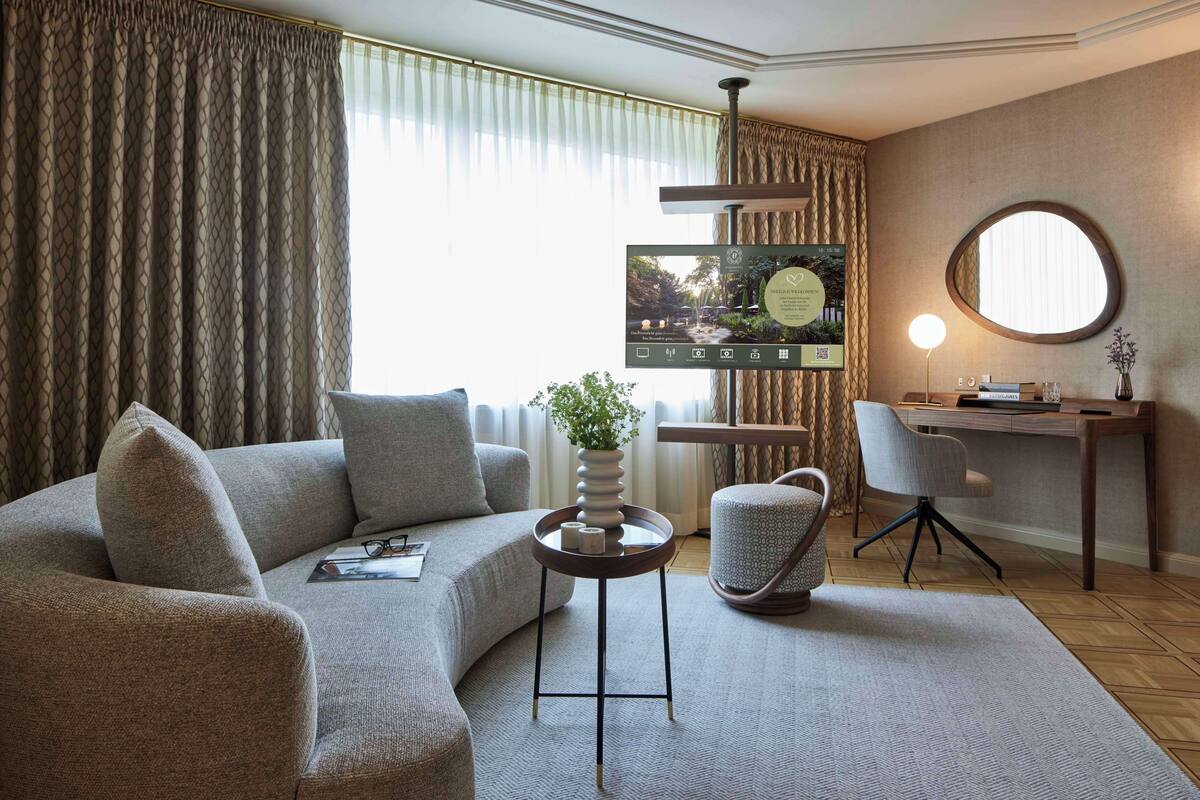 Ein modernes Wohnzimmer mit einer Couch, einem runden Couchtisch, einem Beistelltisch, einem Sessel, einem Schreibtisch mit Computer und einem großen Fenster mit Vorhängen.