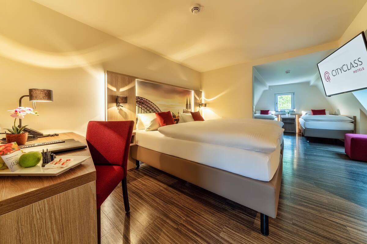 Habitación de hotel con cama, escritorio con una computadora portátil, silla, una mesita con una planta y un espejo.