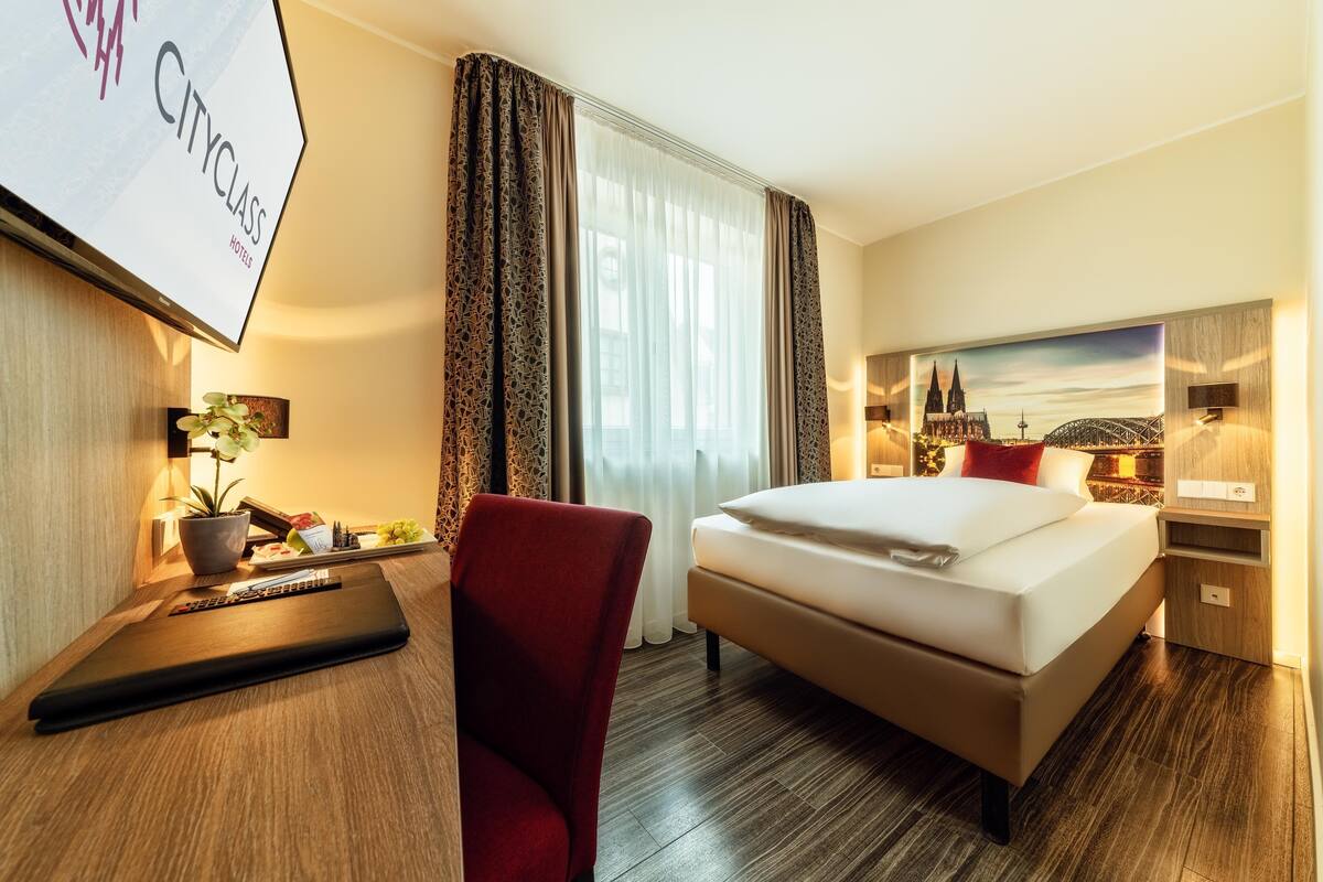 Habitación de hotel con cama, escritorio con una computadora portátil, silla, televisor y un cuadro de un paisaje urbano.