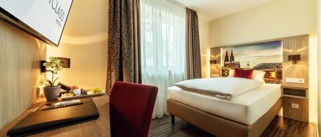 A hotel room with a bed, a desk with a laptop, a chair, a TV screen, and a painting of a cityscape.