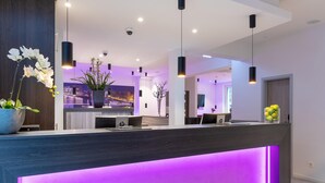 A modern reception area with a dark wood reception desk, purple ambient lighting, and a cityscape view through the window. - CityClass Hotel am Heumarkt (Cologne)