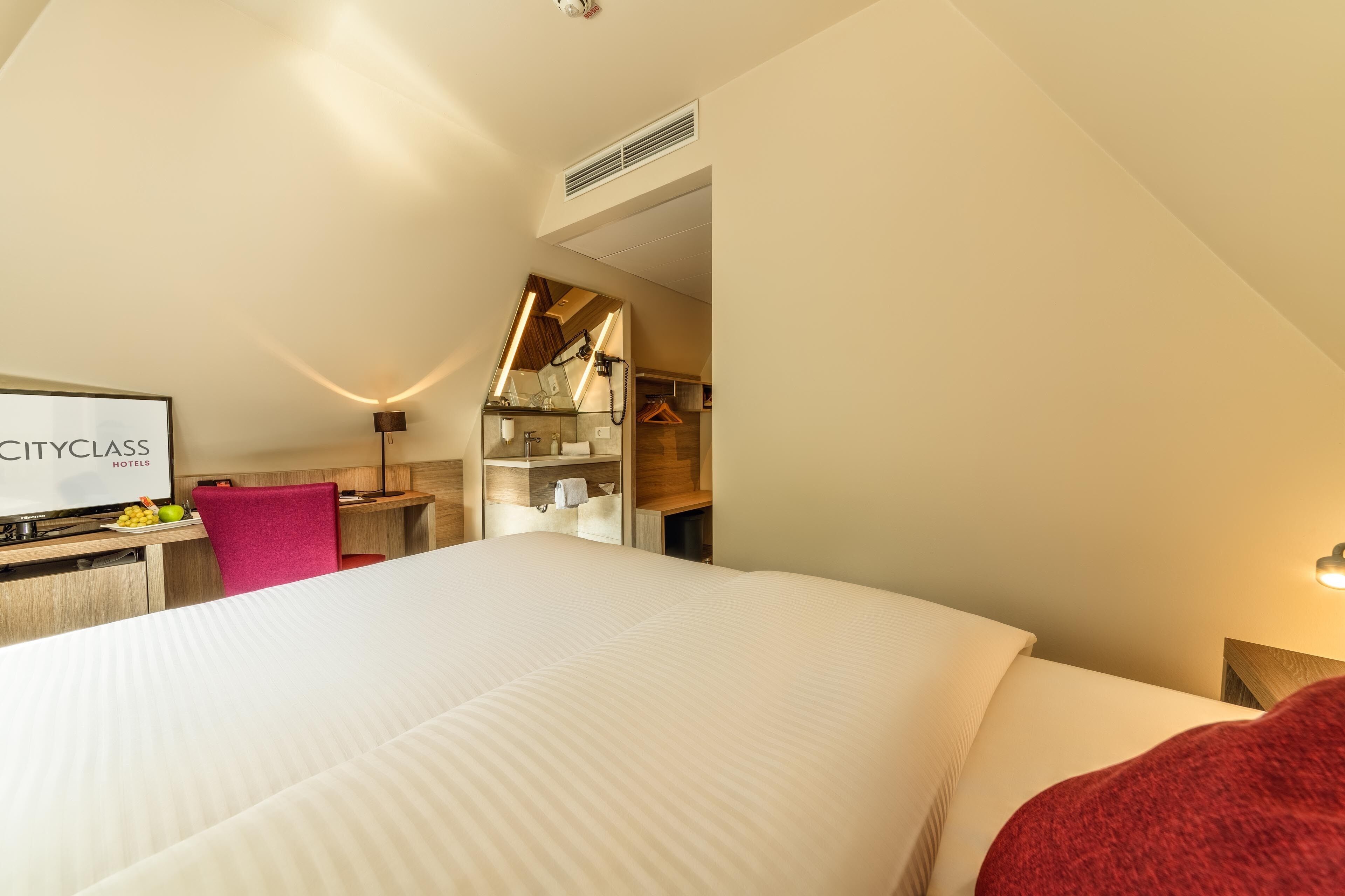 Habitación de hotel con cama, escritorio con silla, espejo y una lámpara de pared.