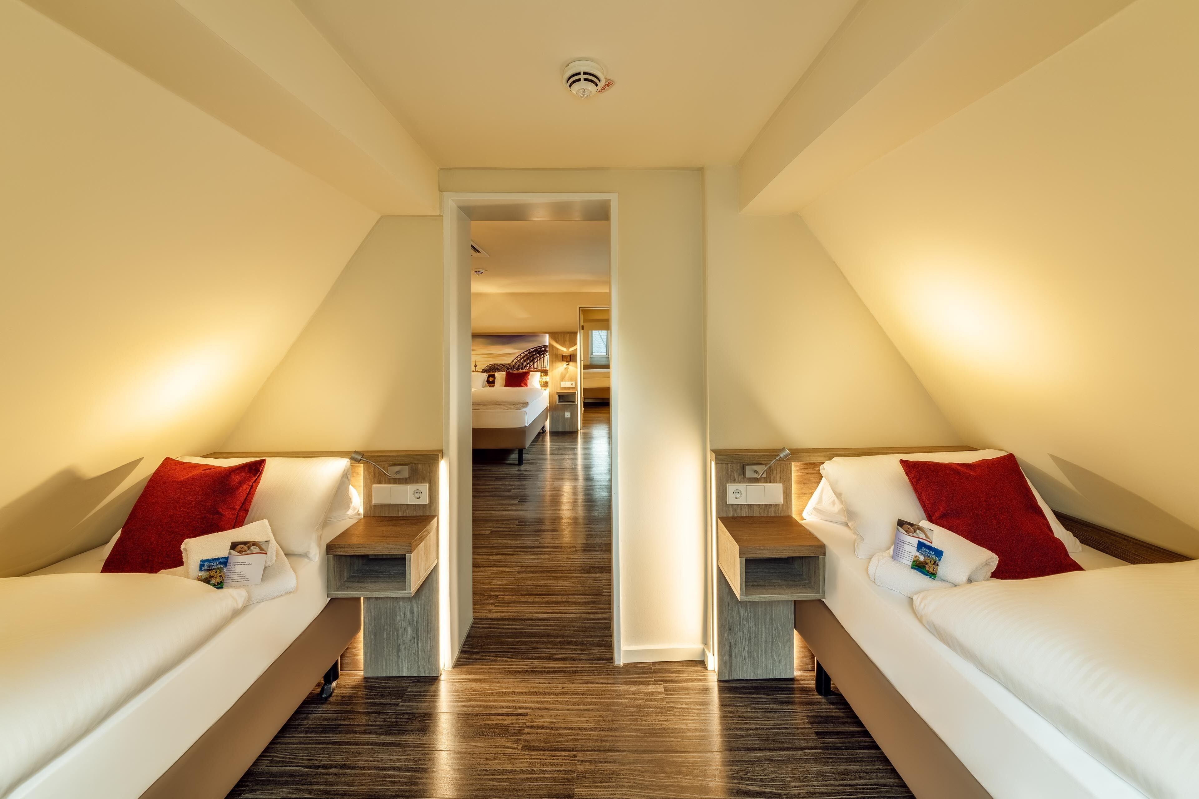 A hotel room with two beds, a wooden floor, a hallway leading to another room, and a ceiling light.
