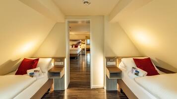 A hotel room with two beds, a wooden floor, a hallway leading to another room, and a ceiling light.